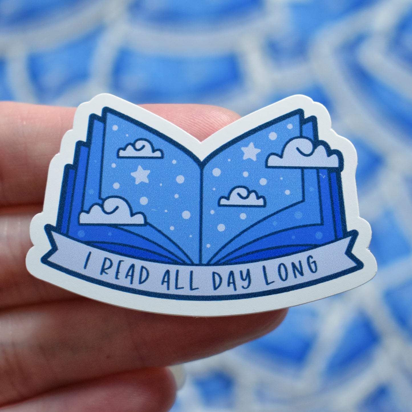 White hand holding sticker. Sticker design is an open book with a mid to dark blue gradient, with off white/pale blue clouds and pale blue dots/stars spread across the design. An off white/pale blue banner at the bottom reads I read all day long. Sticker has a white border. Background is blurry/distance of same stickers.