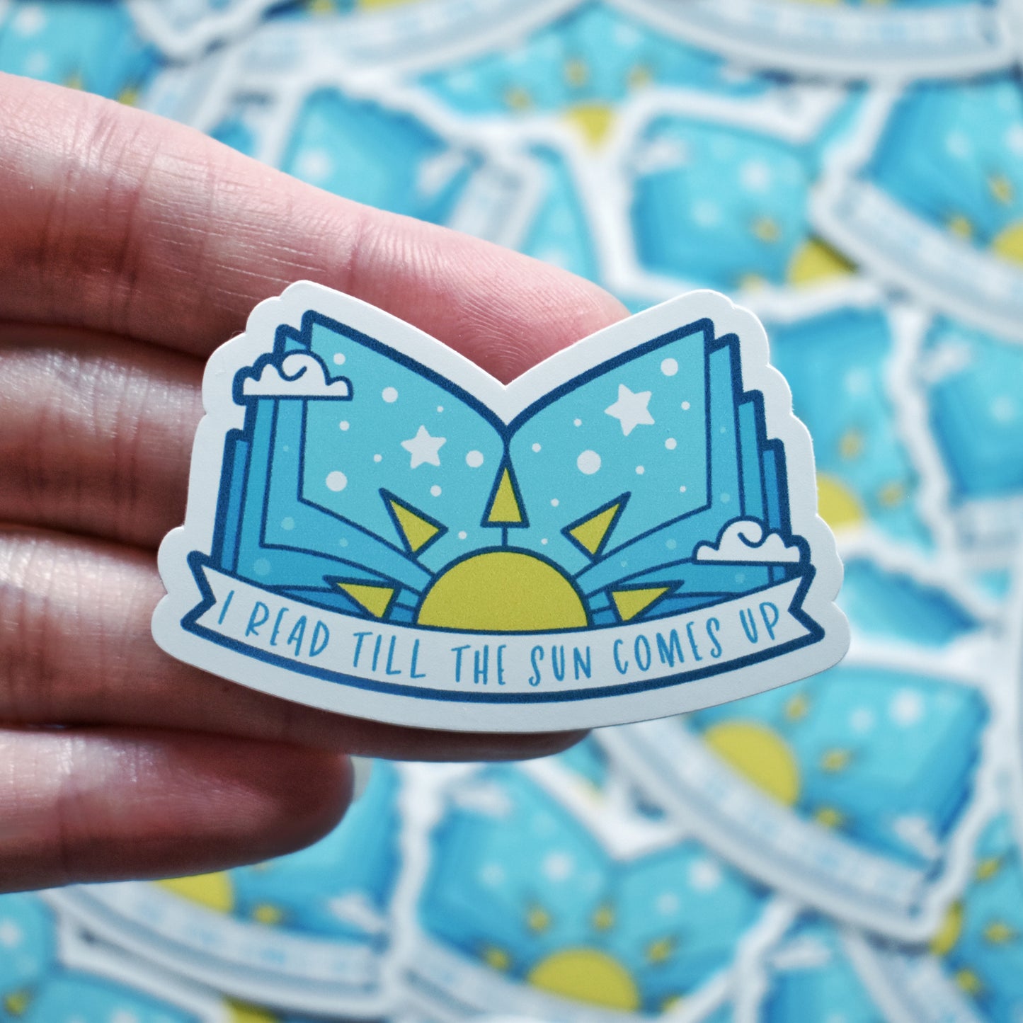 White hand holding sticker. Sticker design is an open book with a pale blue gradient, with a yellow sun in the middle and white dots/stars/clouds spread across the design. A white banner at the bottom reads I read till the sun comes up. Sticker has a white border. Background is blurry/distance of same stickers.