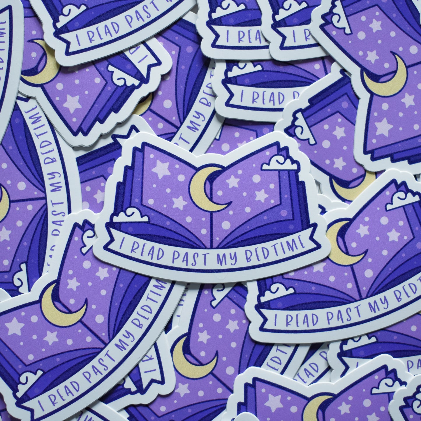Multiple of same sticker scattered. Sticker design is an open book with a purple gradient, with a yellow moon top middle and white dots/stars/clouds spread across the design. A white banner at the bottom reads I read past my bedtime. Sticker has a white border.