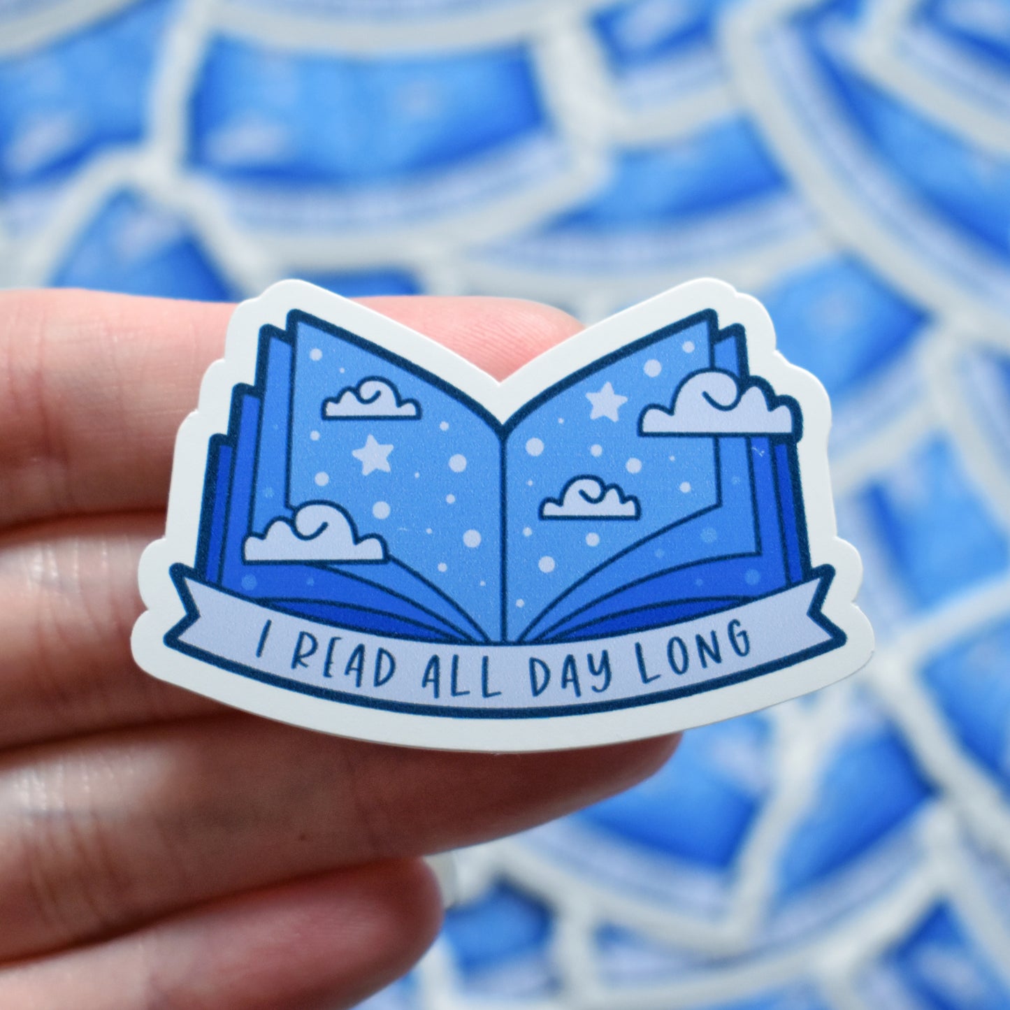 White hand holding sticker. Sticker design is an open book with a mid to dark blue gradient, with off white/pale blue clouds and pale blue dots/stars spread across the design. An off white/pale blue banner at the bottom reads I read all day long. Sticker has a white border. Background is blurry/distance of same stickers.