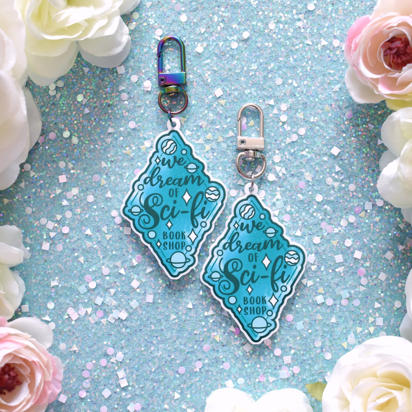 2 acrylic keyrings layed out in the centre of the image. Each featuring a teal diamond design with small planets scattered across the design. Design reads We dream of Sci-fi bookshop. Left keyring is printed on white acrylic with a rainbow clasp. Right keyring is printed on white acrylic with a silver clasp. The background of the image is teal glittery with cream and pink fake flowers around the edge.