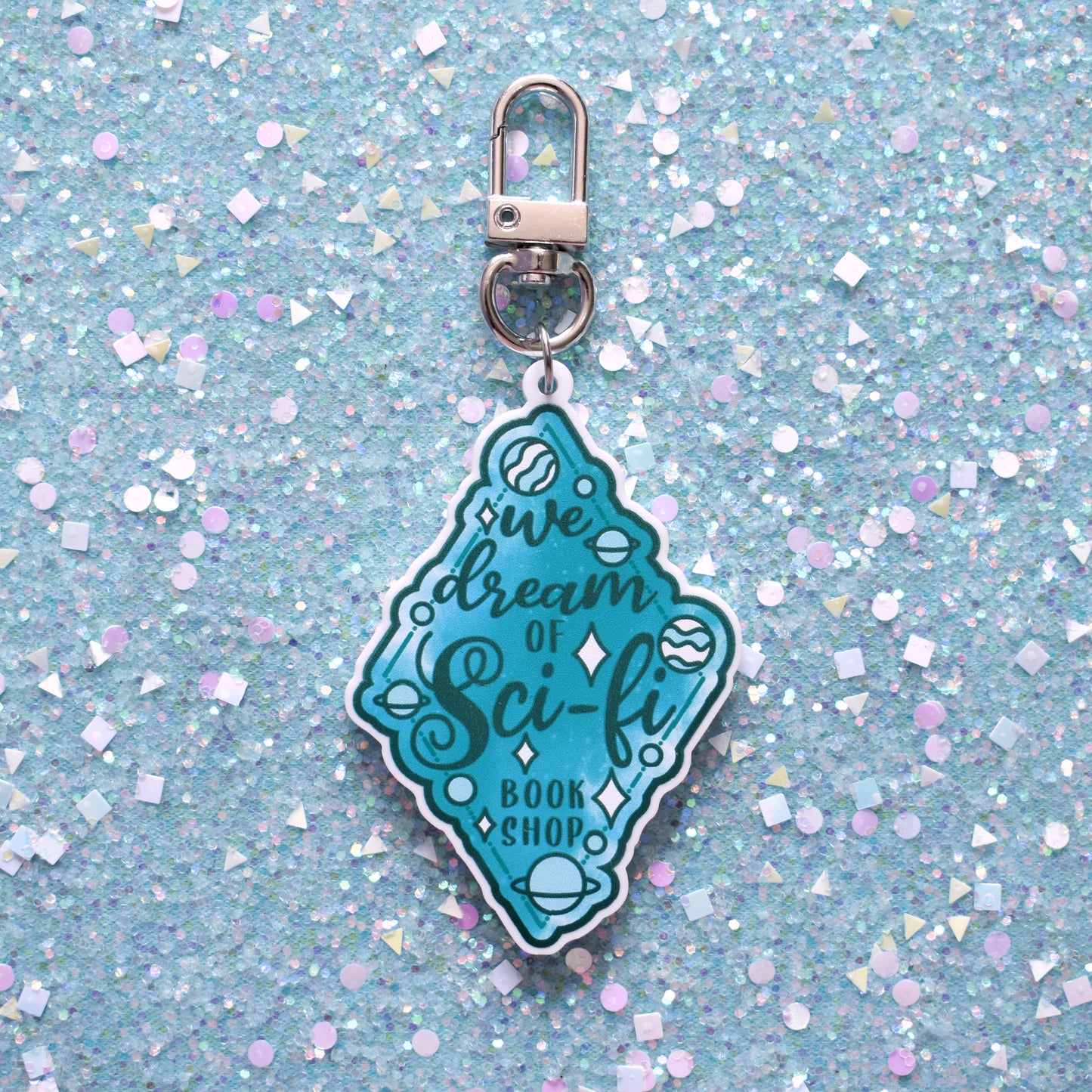 Close up of acrylic keyring in the centre of the image. Featuring a teal diamond design with small planets scattered across the design. Design reads We dream of Sci-fi bookshop. Keyring is printed on white acrylic with a silver clasp. The background of the image is teal glitter.