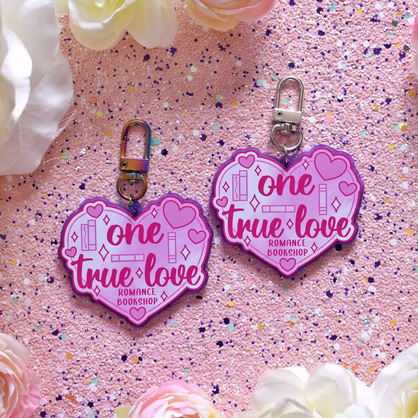 2 acrylic keyrings layed out in the centre of the image. Each featuring a big pink heart with smaller hearts and book spines across the design. Design reads one true love romance bookshop. Left keyring is printed on frosted purple acrylic with a rainbow clasp. Right keyring is printed on frosted purple acrylic with a silver clasp. The background of the image is peach soft sparkly with cream and pink fake flowers around the edge.