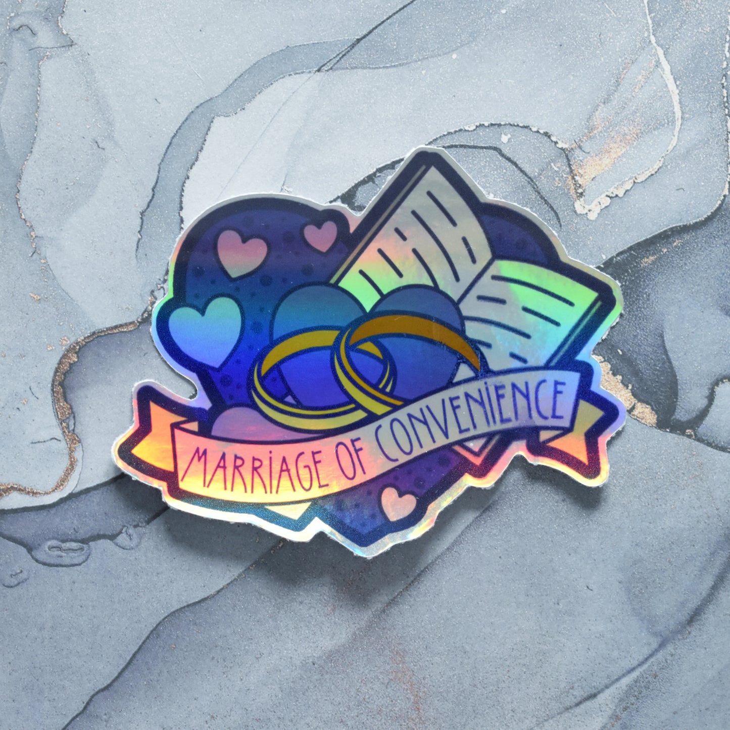 Sticker design is a big blue heart with smaller hearts, an open book and a pair of gold wedding rings inside it. A pale yellow banner across the middle of the design reads marriage of convenience. Sticker has a border all around the design. Holographic effects visible across design. Design has a smear in the print on right side. Background is a grey watercolour style.