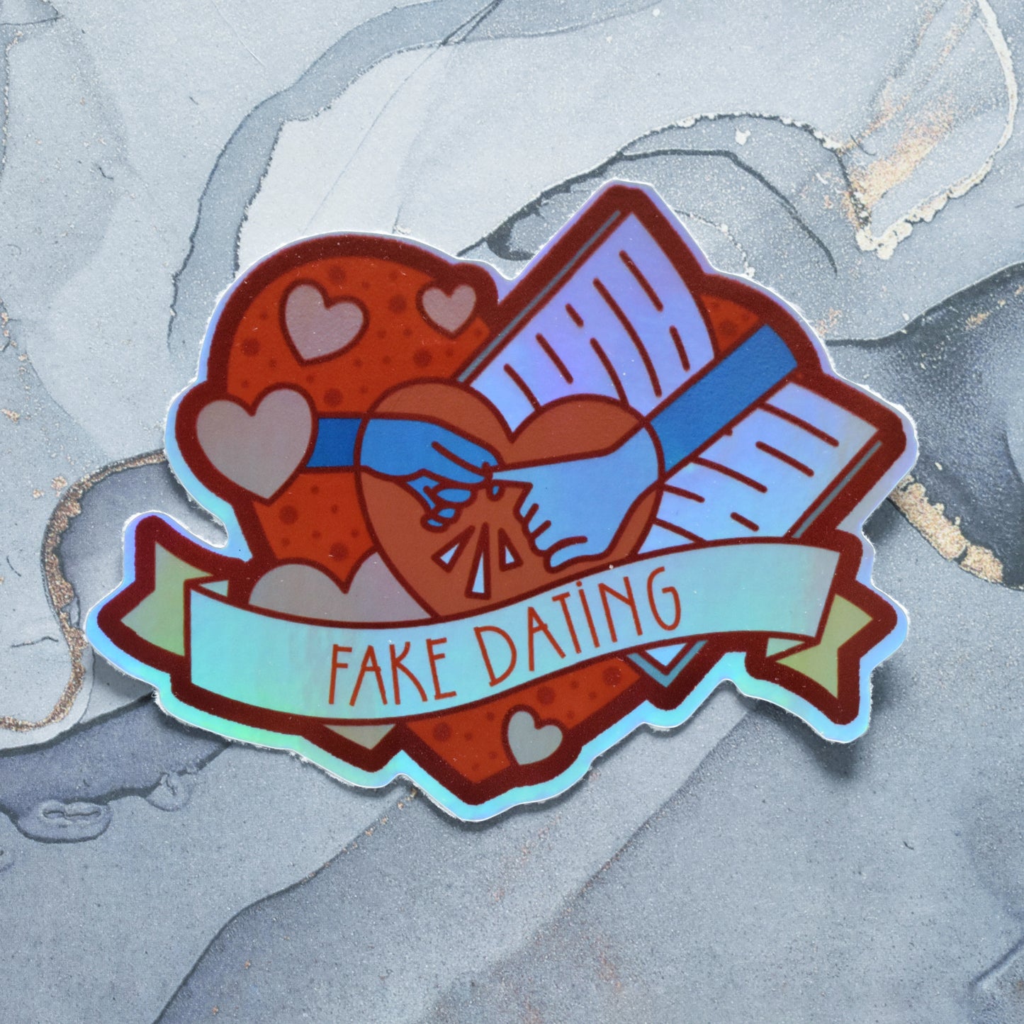 Sticker design is a big red heart with smaller hearts, an open book and a pair of pale grey hands, one hand holding pinky finger of other hand, inside it. A pale yellow banner across the middle of the design reads fake dating. Sticker has a border all around the design. Holographic effects visible across design. Design has uneven border. Background is a grey watercolour style.