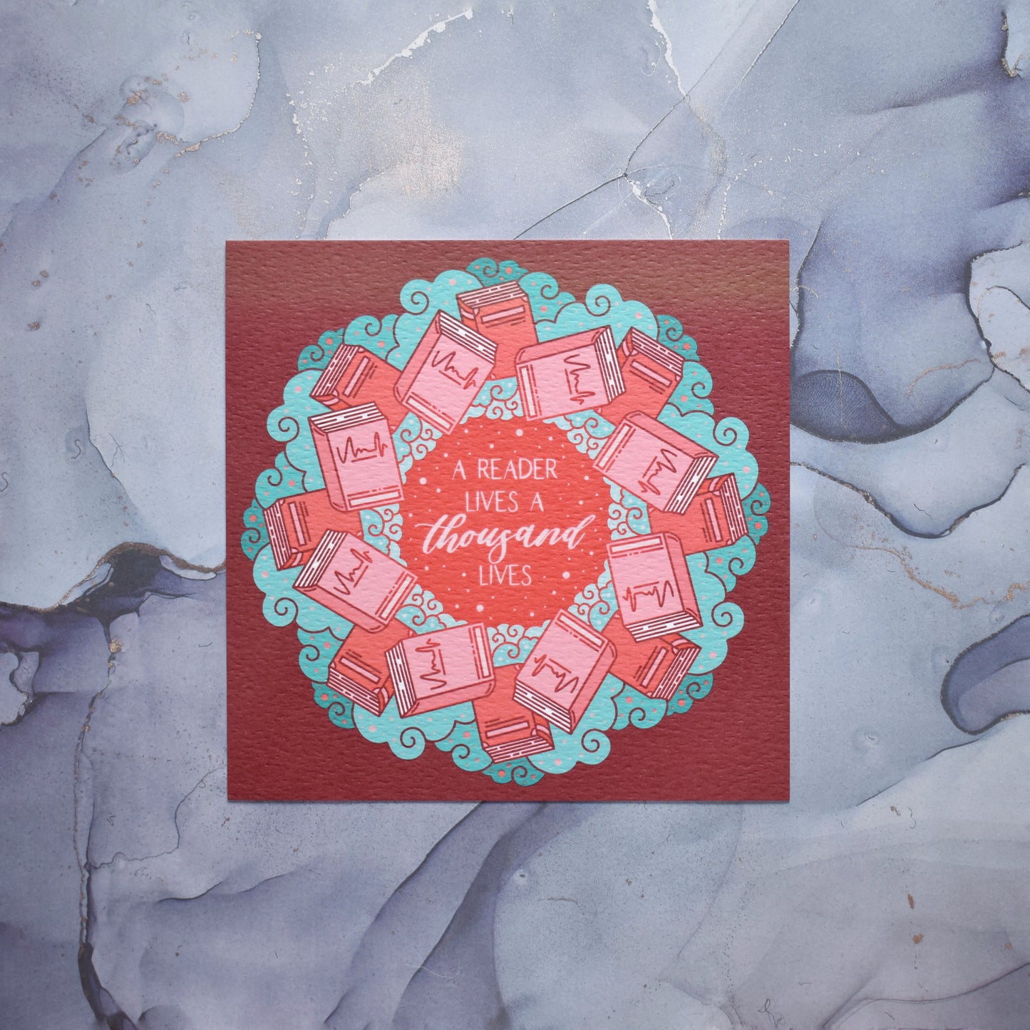 Square print featuring a repeating pattern of red/pink closed books and teal swirls with red around the edge. Band of discolouration across top of print. Design reads a reader lives a thousand lives in pink/red at the centre of the design. Background of photograph is grey watercolour style.