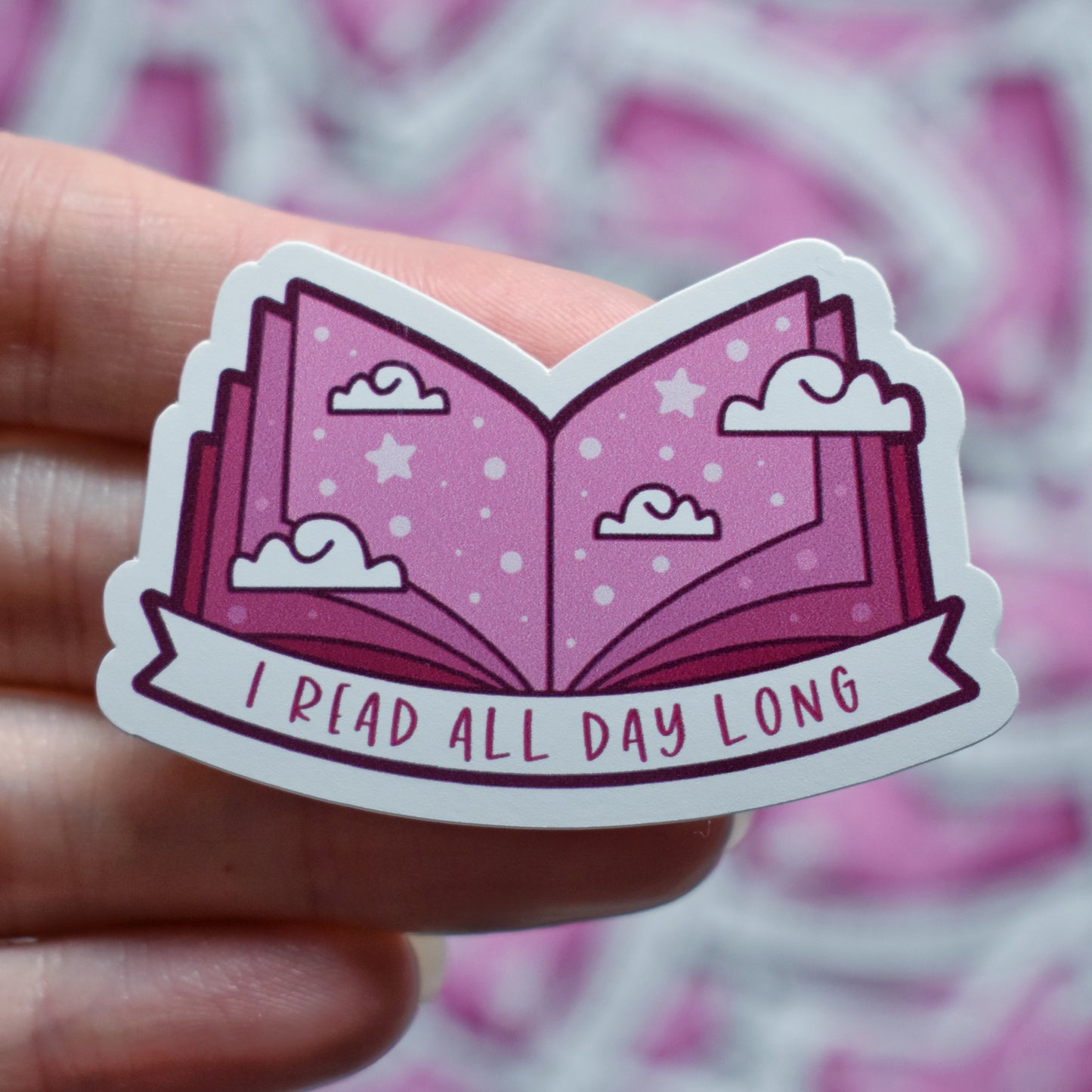 White hand holding sticker. Sticker design is an open book with a mid to dark pink gradient, with white clouds and pale pink dots/stars spread across the design. A white banner at the bottom reads I read all day long. Sticker has a white border. Background is blurry/distance of same stickers.