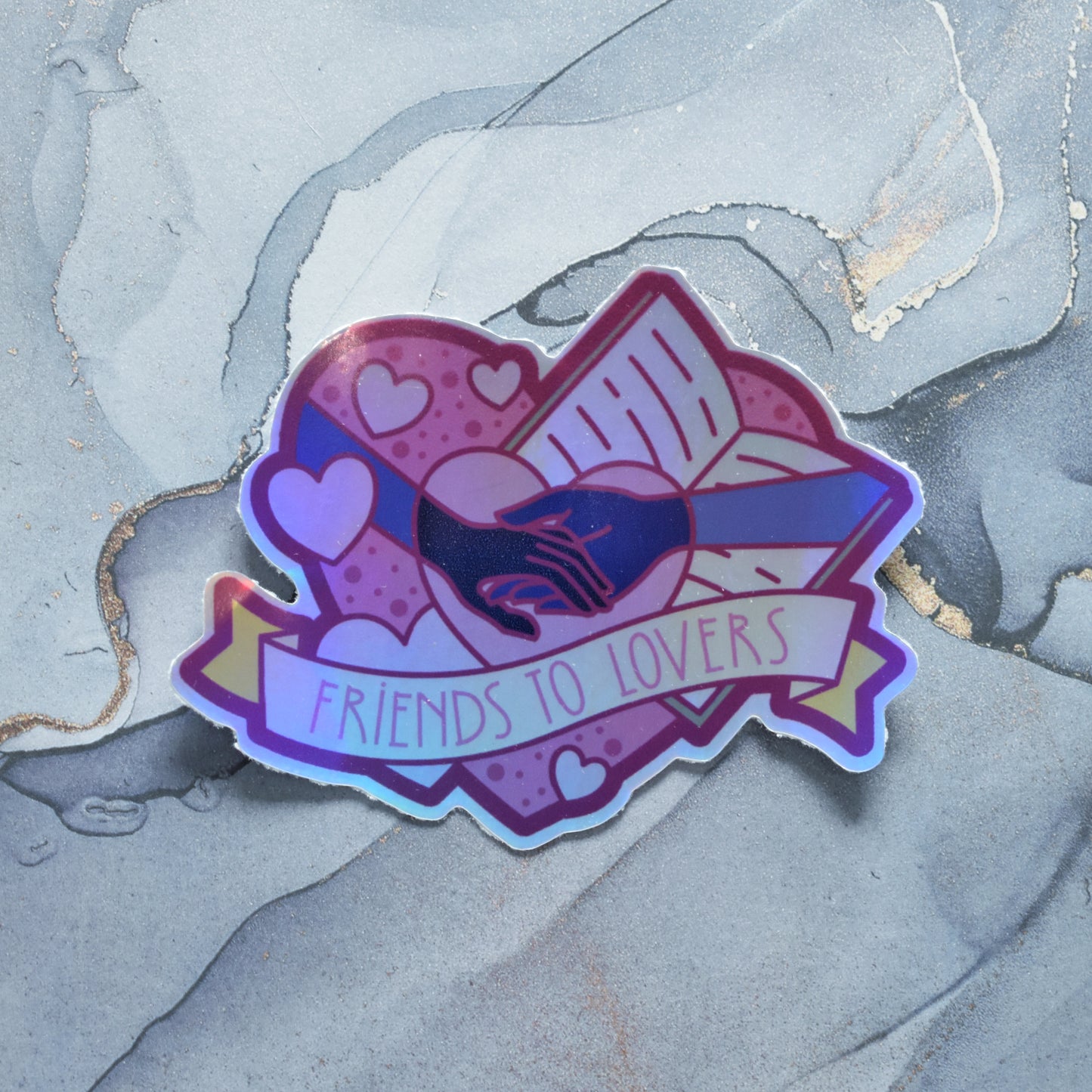 Sticker design is a bigpink heart with smaller hearts, an open book and a pair of grey/blue hands, holding hands, inside it. A pale yellow banner across the middle of the design reads friends to lovers. Sticker has a border all around the design. Holographic effects visible across design. Design has uneven border. Background is a grey watercolour style.