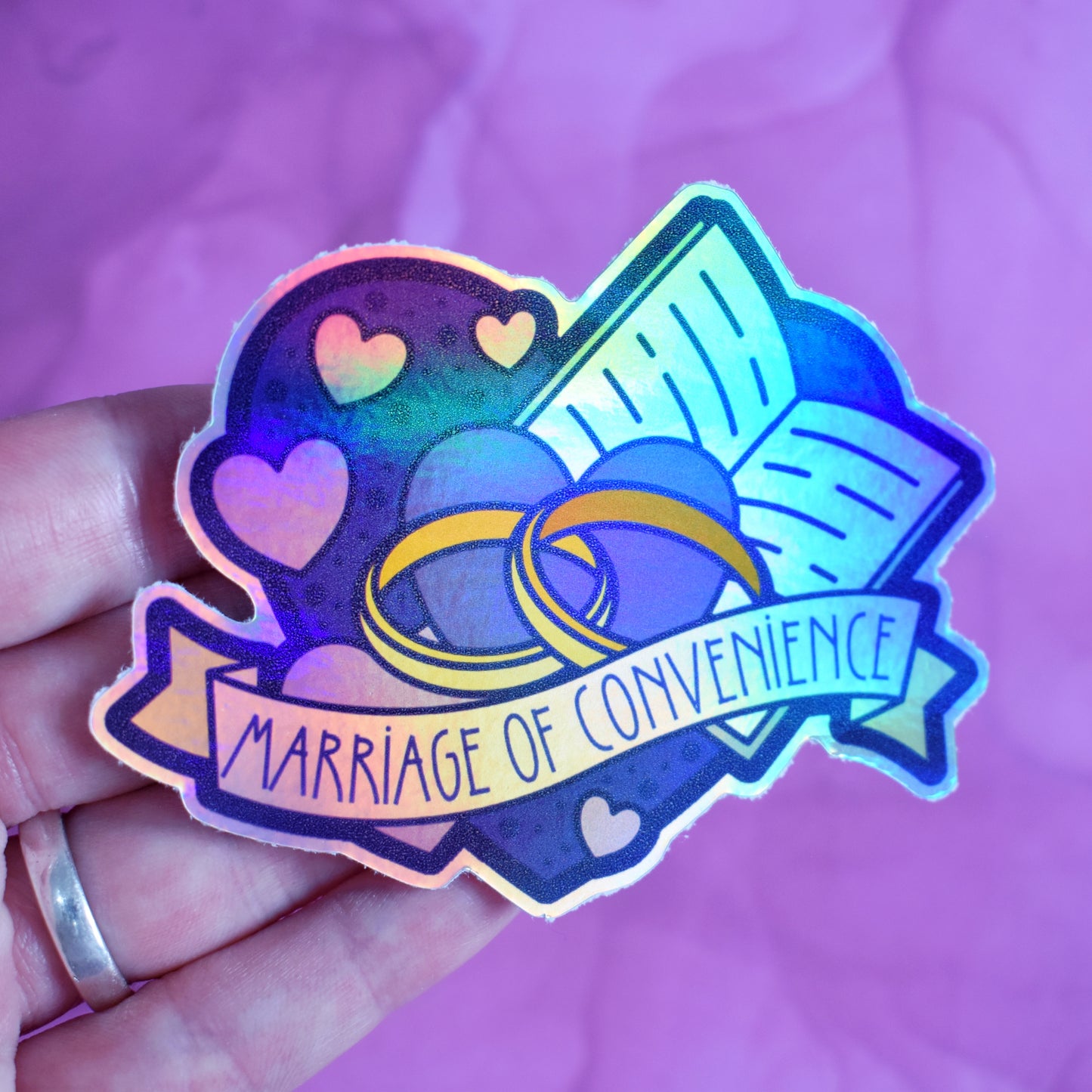 White hand holding sticker, tilted at an angle to show the holographic effect. Sticker design is a big blue heart with smaller hearts, an open book and a pair of gold wedding rings inside it. A pale yellow banner across the middle of the design reads marriage of convenience. Sticker has a border all around the design. Background is blurry/distance of a pinkish watercolour style.