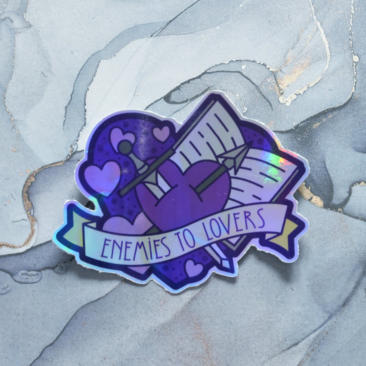Sticker design is a big purple heart with smaller hearts, an open book and a sword and arrow crossed inside it. A pale yellow banner across the middle of the design reads enemies to lovers. Sticker has a border all around the design. Holographic effects visible across design. Design has uneven border and a crease on left side. Background is a grey watercolour style.
