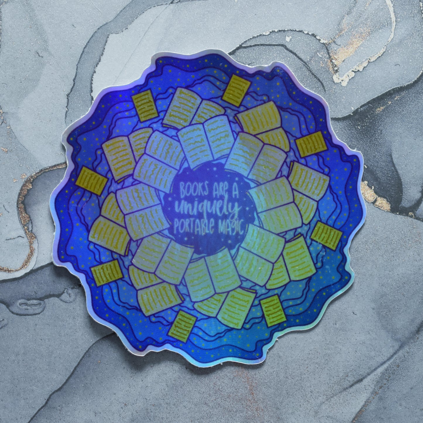 Holographic sticker, featuring a slightly blurry repeating pattern of yellow open books and blue swirls with purple around the edge. Design reads books are a uniquely portable magic in yellow at the centre of the design. Background is grey watercolour style.