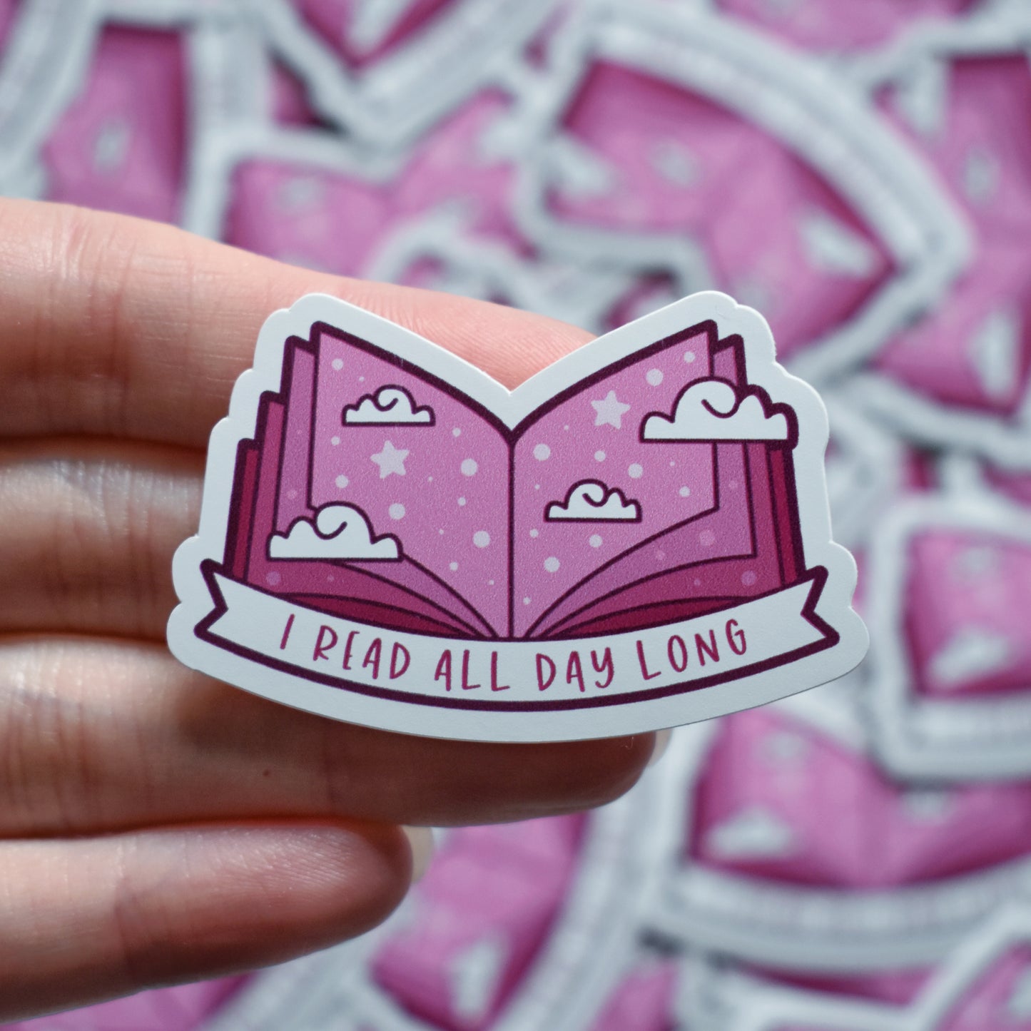White hand holding sticker. Sticker design is an open book with a mid to dark pink gradient, with white clouds and pale pink dots/stars spread across the design. A white banner at the bottom reads I read all day long. Sticker has a white border. Background is blurry/distance of same stickers.