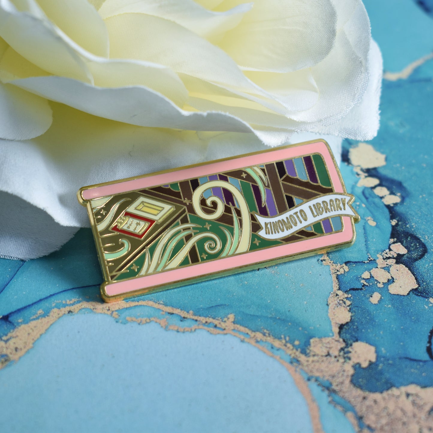 Gold metal enamel pin in centre of image. Enamel pin is shaped like the end of a book in light pink and features a scene in a library. Shelves of books on the right with a desk on the left. Desk has an open clow book and swirls of white and green with sparkles. A small banner reads kinomoto library on the bottom right of the design. Background of photograph is blue water inspired with a fake cream flower.