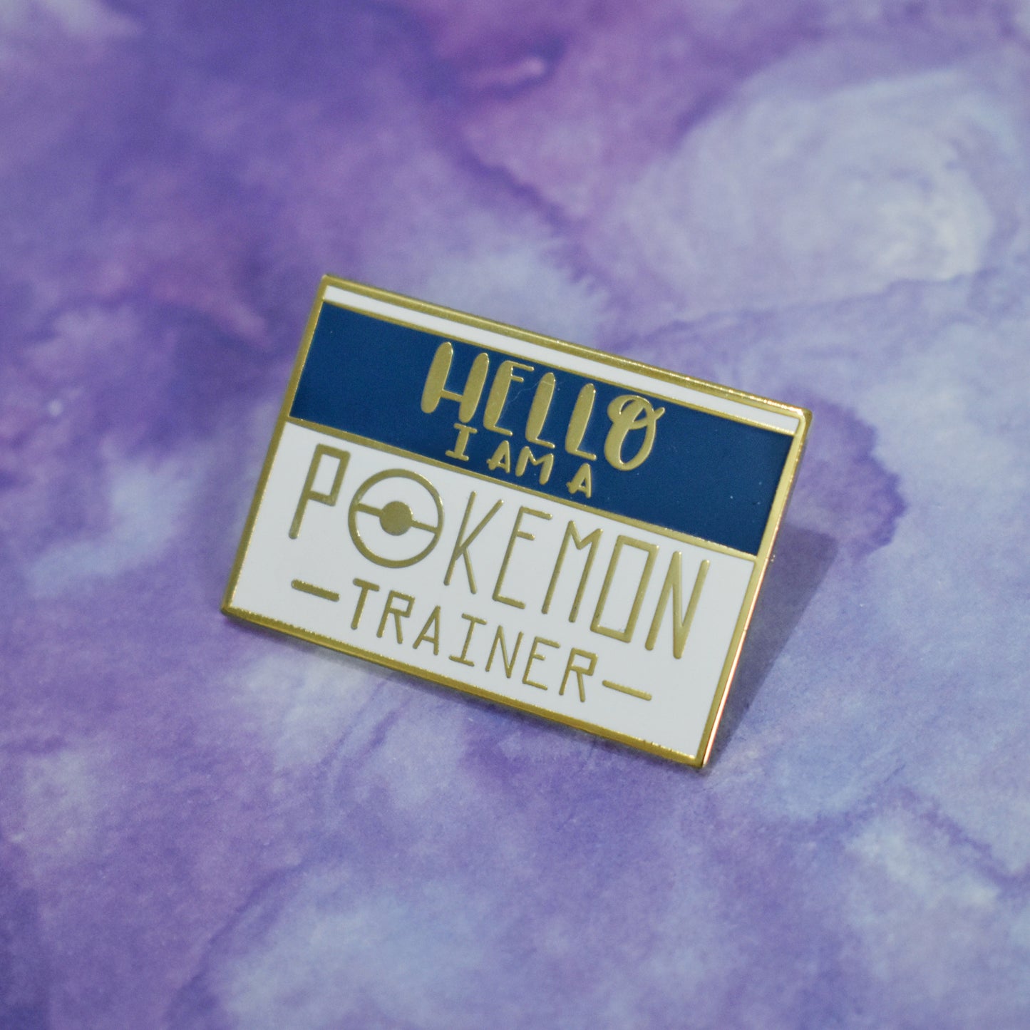Rectangle gold metal enamel pin at an angle in center of photo. White background with a blue band across most of top half. Hello I am a pokemon trainer written across design. Pokemon trainer in a large skinny sans serif style font. Background of photo is purple watercolour style.