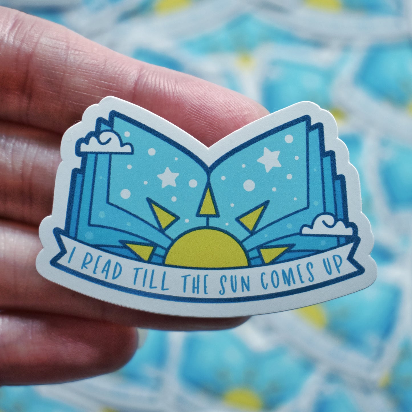 White hand holding sticker. Sticker design is an open book with a pale blue gradient, with a yellow sun in the middle and white dots/stars/clouds spread across the design. A white banner at the bottom reads I read till the sun comes up. Sticker has a white border. Background is blurry/distance of same stickers.