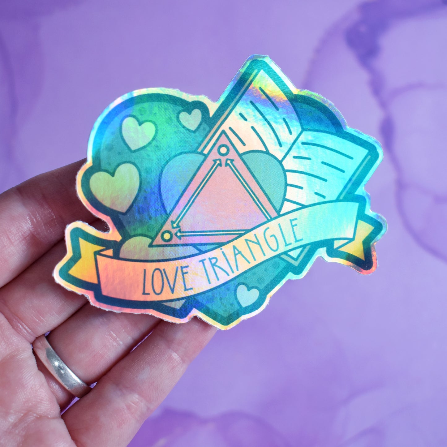 White hand holding sticker, tilted at an angle to show the holographic effect. Sticker design is a big green/teal heart with smaller hearts, an open book and a pink triangle with arrows pointing to each corner inside it. A pale yellow banner across the middle of the design reads love triangle.. Sticker has a border all around the design. Background is blurry/distance of a pinkish purple watercolour style.