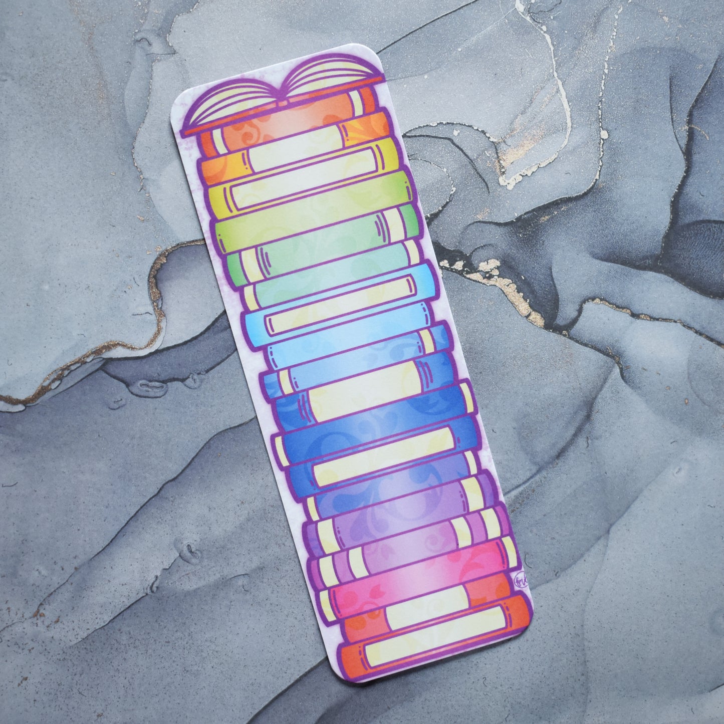 White hand holding bookmark. Bookmark with rounded edges featuring a rainbow bookstack with an open book at the top of the design. Design is slightly off centre on the bookmark. Background is grey watercolour style.