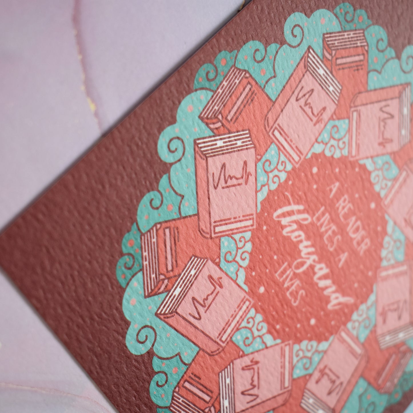Close up of corner of design to show the texture of the print which is slightly raised. Square print featuring a repeating pattern of red/pink closed books and teal swirls with red around the edge. Design reads a reader lives a thousand lives in pink/red at the centre of the design. Background of photograph is pink/blue watercolour style.