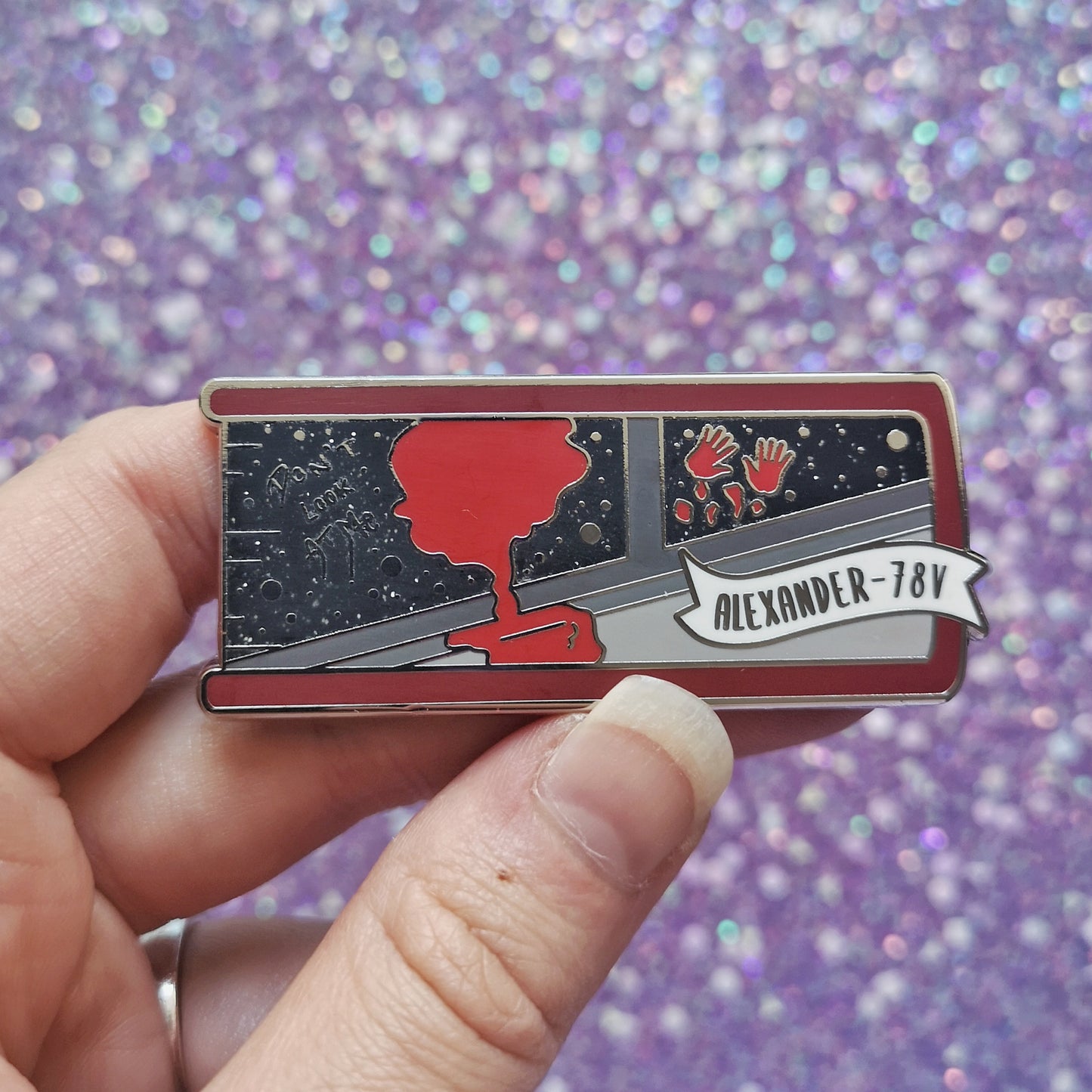 White hand holding silver metal enamel pin. Enamel pin is shaped like the end of a book in red and features a scene with the inside of a grey spaceship looking out into a black glittery space. With a splash of red dripping down and 2 red handprints. A small banner reads alexander-78V on the bottom right of the design. Background of photograph is sparkly purple.