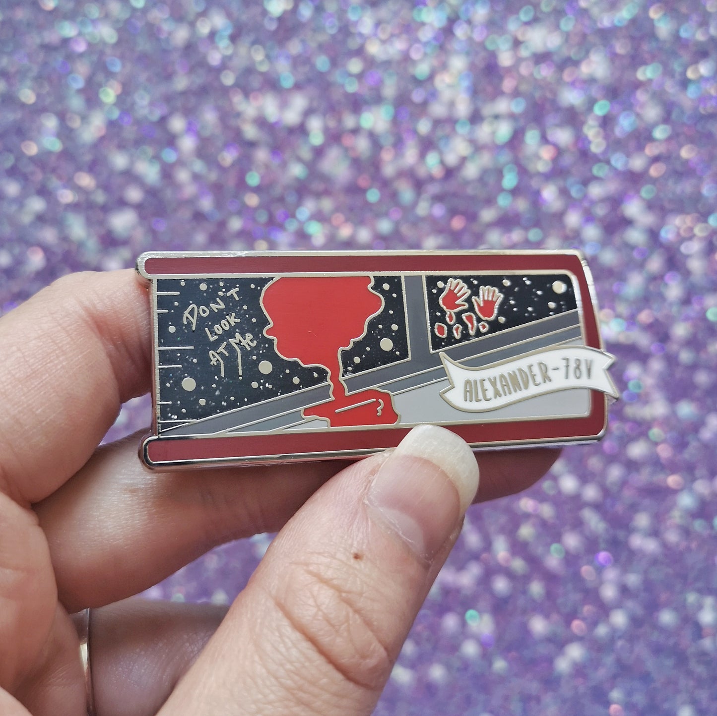 White hand holding enamel pin, tilted to make the silver metal catch more light. Enamel pin is shaped like the end of a book in red and features a scene with the inside of a grey spaceship looking out into a black glittery space. With a splash of red dripping down and 2 red handprints. A small banner reads alexander-78V on the bottom right of the design. Background of photograph is sparkly purple.