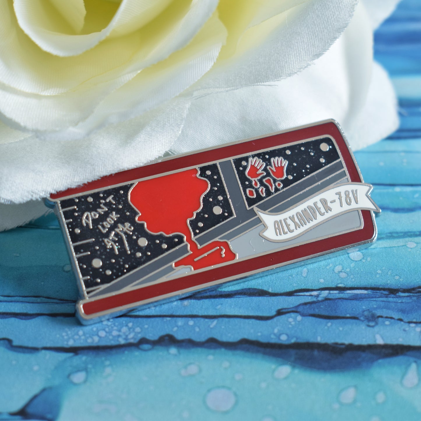Silver metal enamel pin in centre of image. Enamel pin is shaped like the end of a book in red and features a scene with the inside of a grey spaceship looking out into a black glittery space. With a splash of red dripping down and 2 red handprints. A small banner reads alexander-78V on the bottom right of the design. Background of photograph is blue water inspired with a fake white flower.