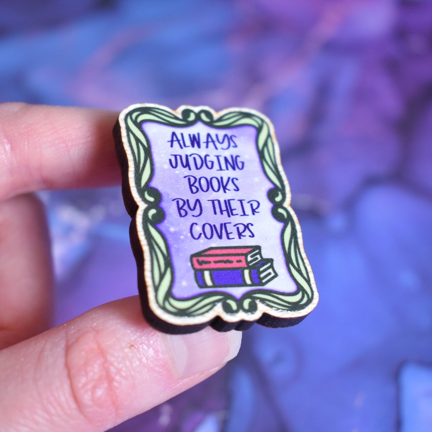 White hand holding small rectangular wooden pin in centre of photo at an angle, which shows the cut edge of the pin which is darker than the wood visable on the front. Design features a swirly green frame with a purple centre. In the centre is written always judging books by their covers with 2 books underneath. Wooden pin has a gloss so has a little shiny around the edge. Background is soft watercolour blue and purple style.