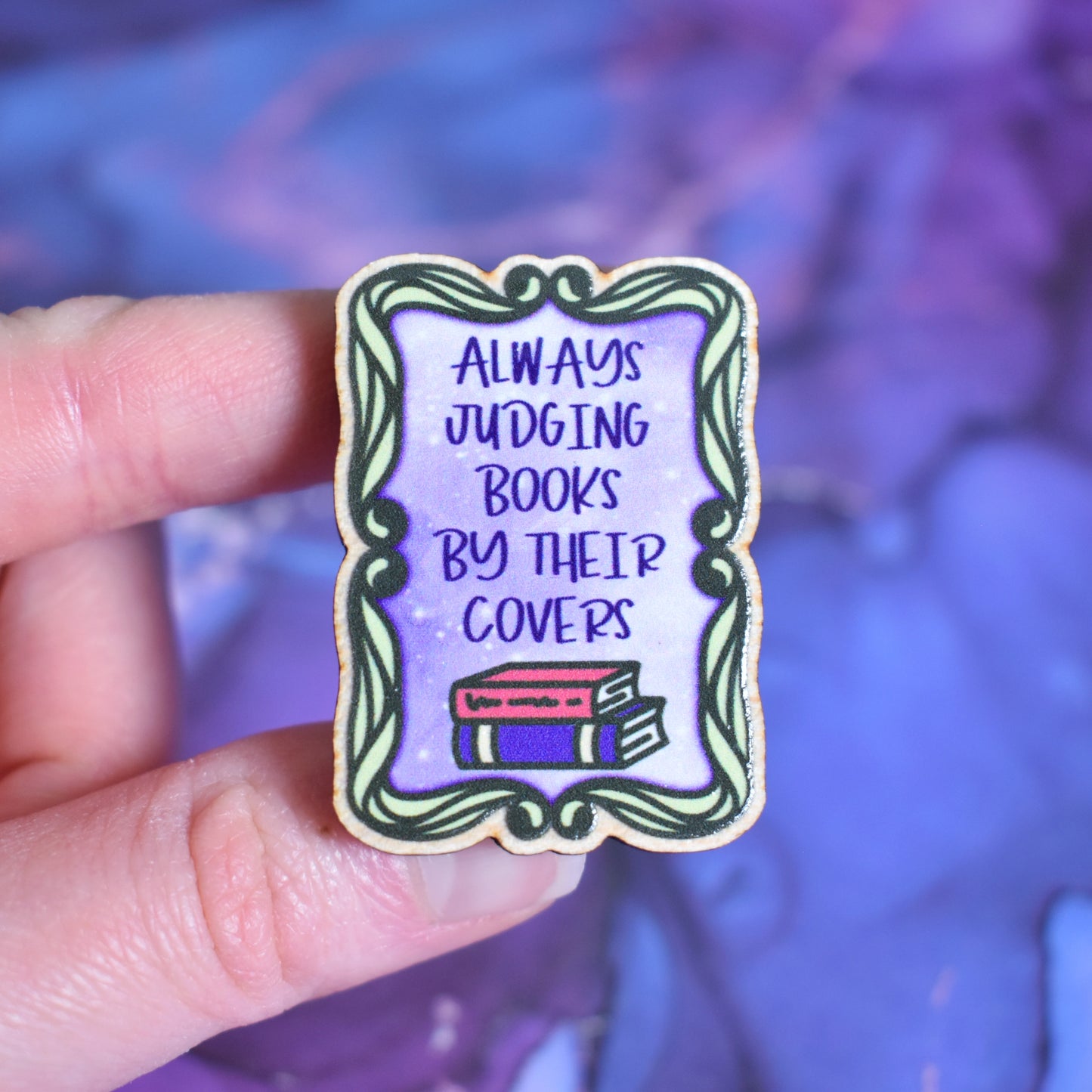 White hand holding small rectangular wooden pin in centre of photo. Design features a swirly green frame with a purple centre. In the centre is written always judging books by their covers with 2 books underneath. Wooden pin has a gloss so has a little shiny around the edge. Background is soft watercolour blue and purple style.