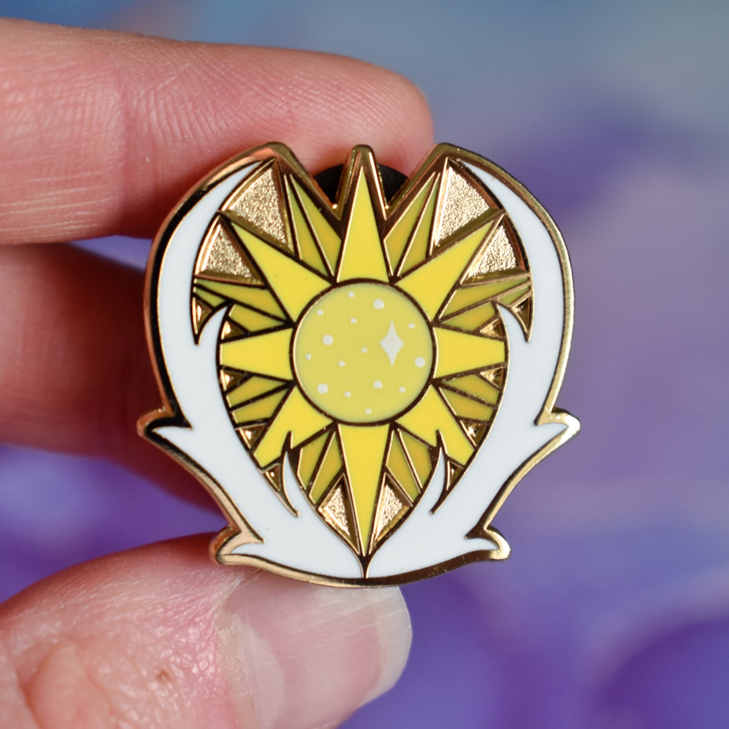 White hand holding gold metal enamel pin. Enamel pin has a yellow many pointed sun at its centre with sparkles and is framed on the bottom and either side by a pair of white antlers. Background of photograph is blurry purple/blue coloured.