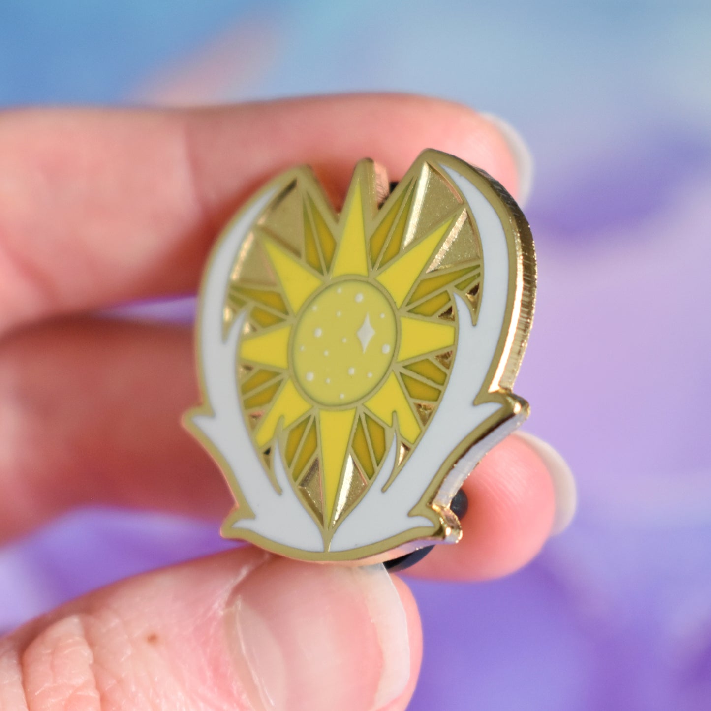 White hand holding gold metal enamel pin tilted to make the metal catch more light. Enamel pin has a yellow many pointed sun at its centre with sparkles and is framed on the bottom and either side by a pair of white antlers. Background of photograph is blurry purple/blue coloured.