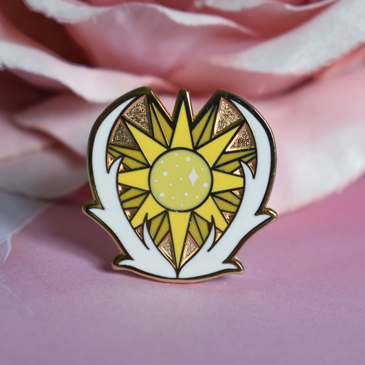 Gold metal enamel pin in centre of image. Enamel pin has a yellow many pointed sun at its centre with sparkles and is framed on the bottom and either side by a pair of white antlers. Background of photograph is pink water inspired with a fake dusty pink flower.
