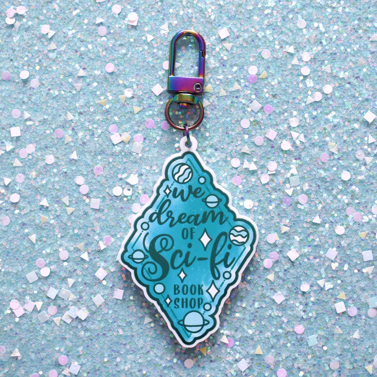 Close up of acrylic keyring in the centre of the image. Featuring a teal diamond design with small planets scattered across the design. Design reads We dream of Sci-fi bookshop. Keyring is printed on white acrylic with a rainbow clasp. The background of the image is teal glitter.
