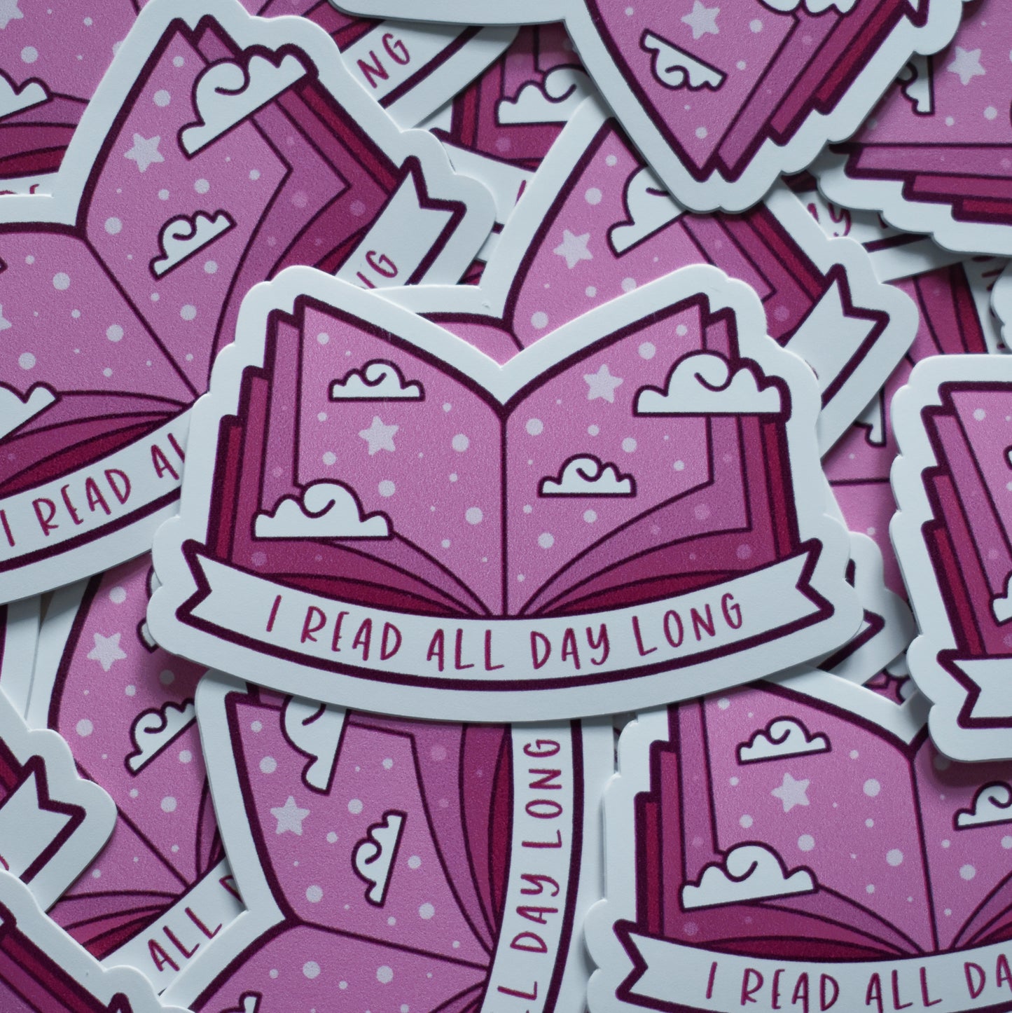 Multiple of same sticker scattered. Sticker design is an open book with a mid to dark pink gradient, with white clouds and pale pink dots/stars spread across the design. A white banner at the bottom reads I read all day long. Sticker has a white border. Background is blurry/distance of same stickers.