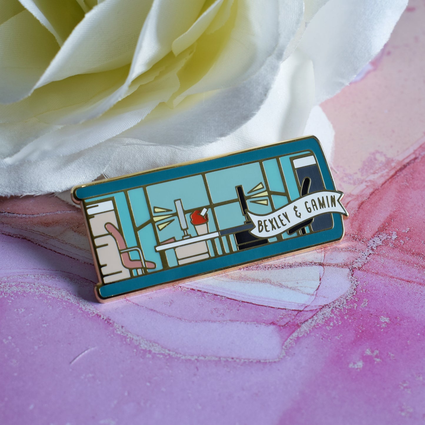 Gold metal enamel pin in centre of image. Enamel pin is shaped like the end of a book in teal and features an office scene in teal, peach and black. 2 office chairs either side of a shared desk in the centre. A small banner reads bexley & gamin on the bottom right of the design. Background of photograph is pinky/purple watercolour style with a fake white flower.