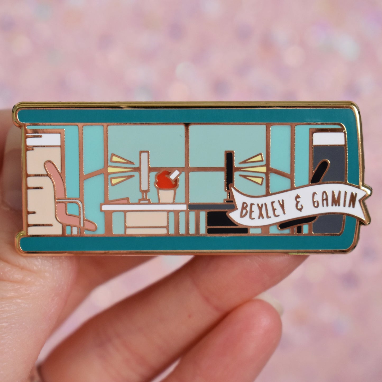 White hand holding gold metal enamel pin. Enamel pin is shaped like the end of a book in teal and features an office scene in teal, peach and black. 2 office chairs either side of a shared desk in the centre. A small banner reads bexley & gamin on the bottom right of the design. Background of photograph is peachy coloured.