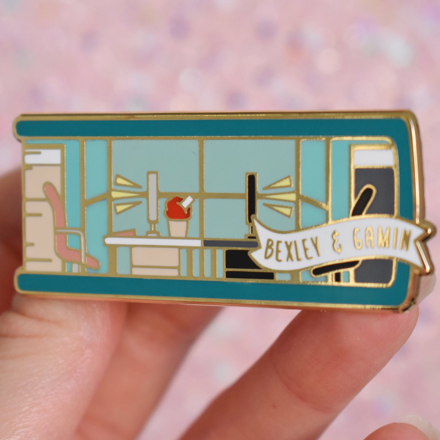 White hand holding gold metal enamel pin tilted to make the metal catch more light. Enamel pin is shaped like the end of a book in teal and features an office scene in teal, peach and black. 2 office chairs either side of a shared desk in the centre. A small banner reads bexley & gamin on the bottom right of the design. Background of photograph is peachy coloured.