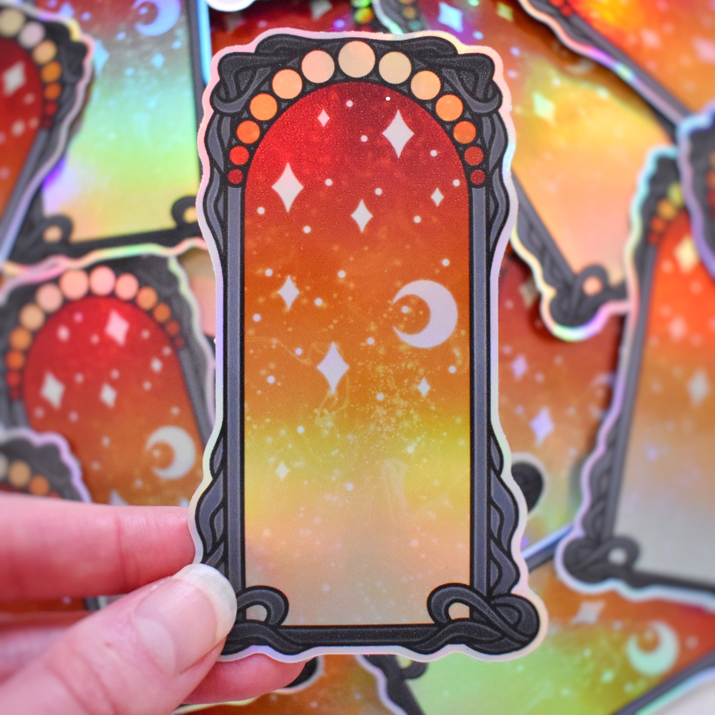 White hand holding sticker. Sticker design is a black curly frame with a gradient of circles across the top, from yellowish/peach to red. Centre of the design is a yellowish/peach to red gradient with white/hazy stars and moon. Background is blurry/distance of same stickers.