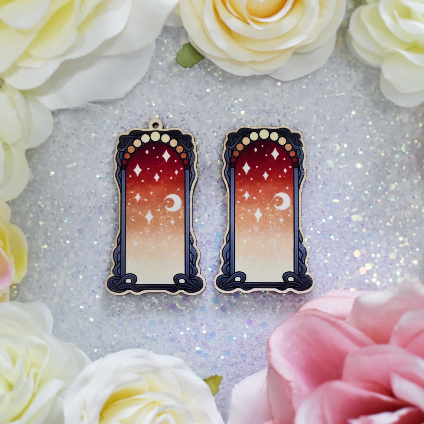 2 wooden charms, 1 with a loop at the top, 1 without next to each other on a sparkly background surrounded by fake flowers. Designs have black detailed frame. Gradient colour yellow through to red with stars and a moon inside frame. Fake flowers in white, yellow and pink are visible around edge of photo.