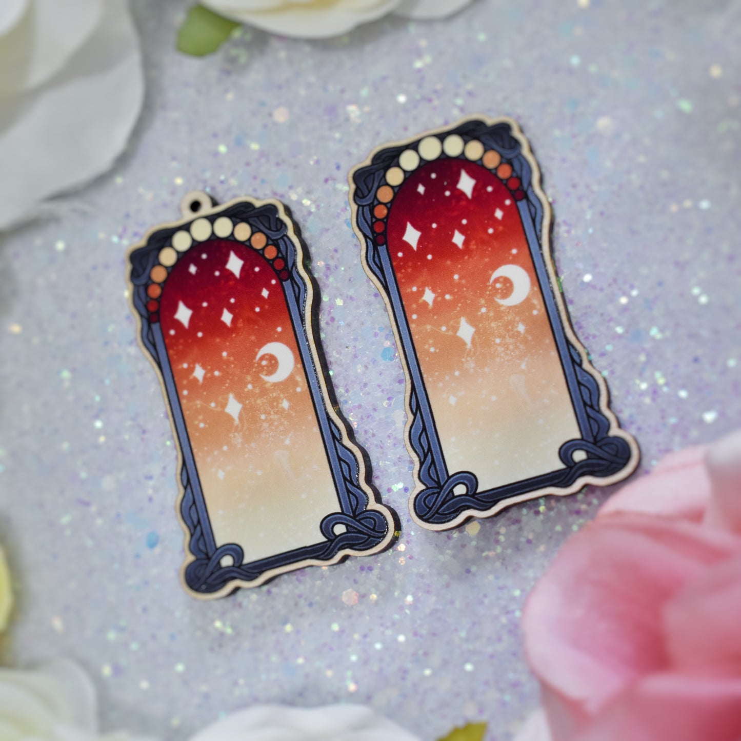 2 wooden charms, 1 with a loop at the top, 1 without next to each other at an angle on a sparkly background. Designs have black detailed frame. Gradient colour yellow through to red with stars and a moon inside frame. Fake flowers in white, yellow and pink are visible around edge of photo.