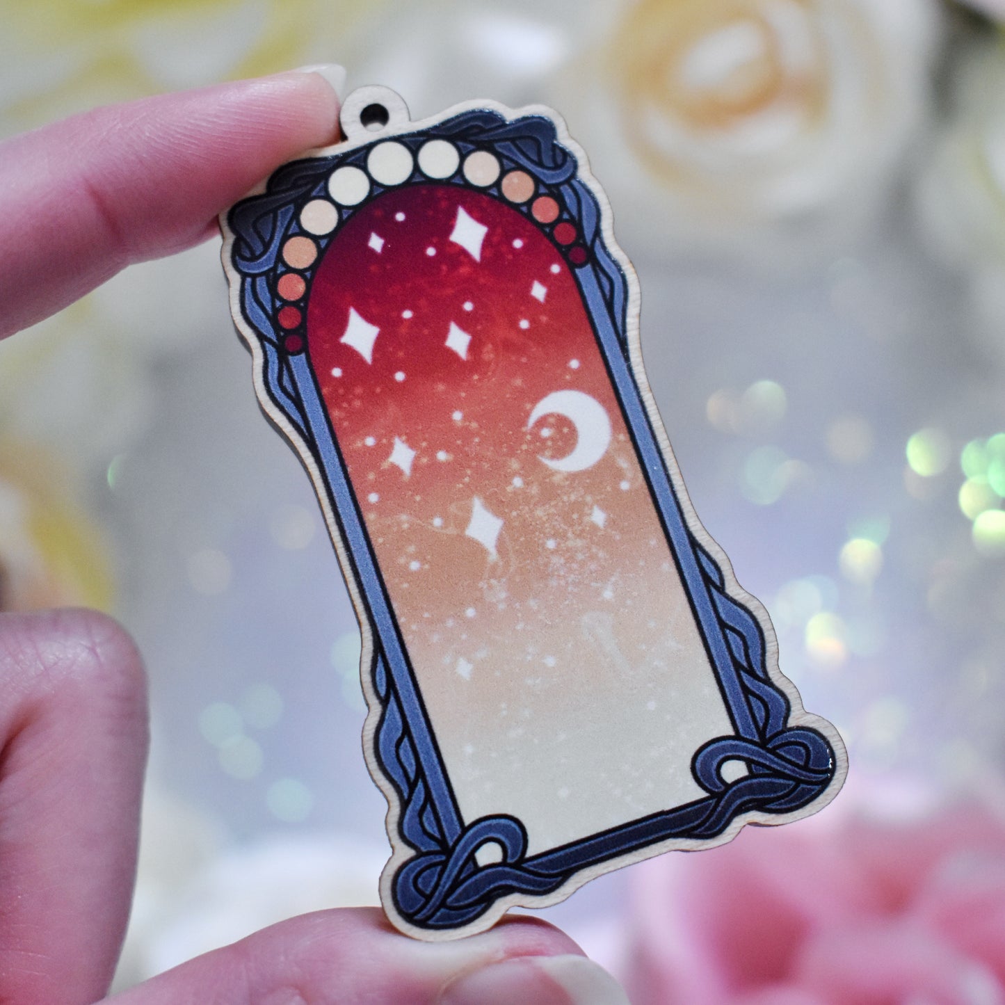 White hand holding wooden charm with top loop. Design has a black detailed frame. Gradient colour yellow through to red with stars and a moon inside frame. Blurred fake flowers in white, yellow and pink are visible around edge of photo.