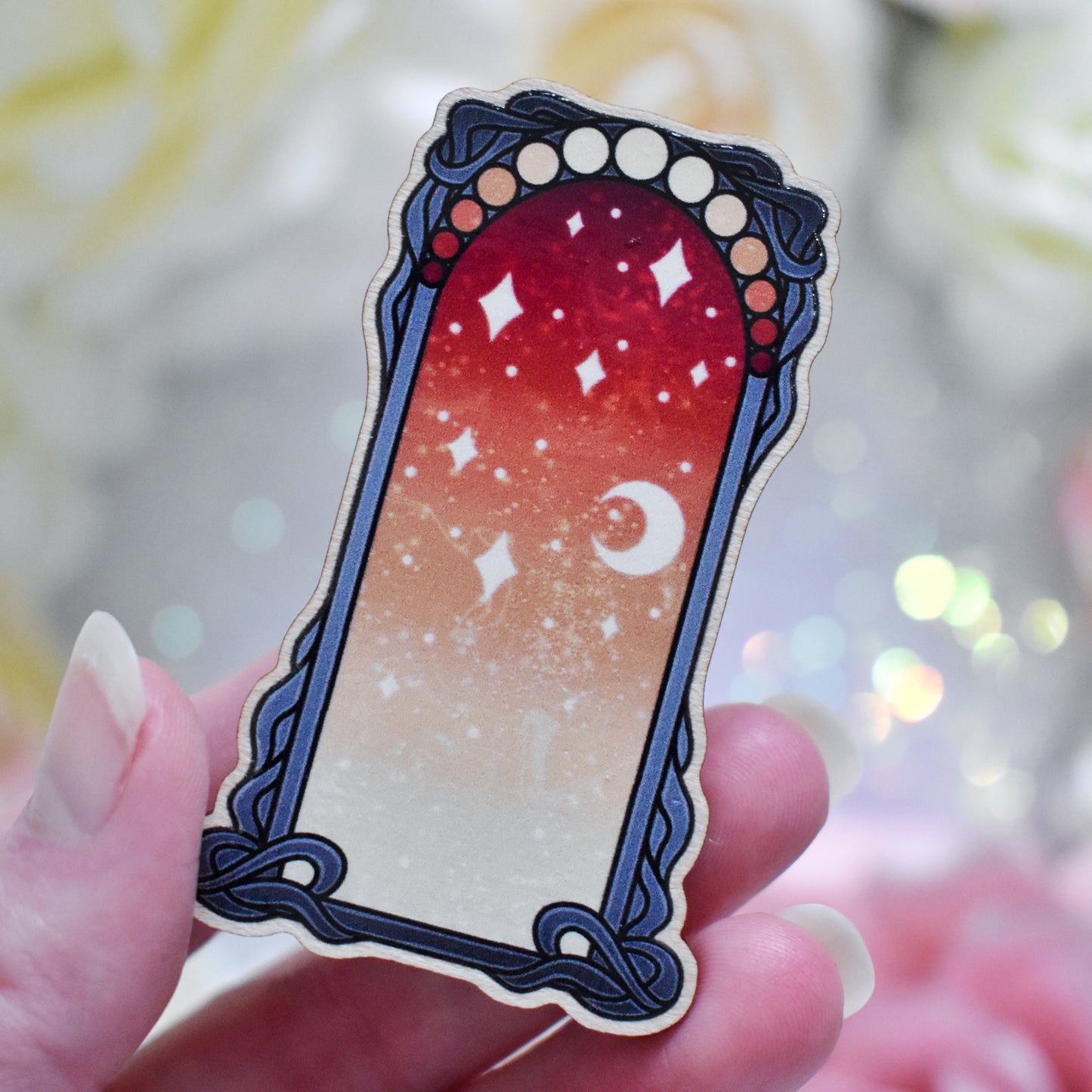 White hand holding wooden charm without top loop. Design has a black detailed frame. Gradient colour yellow through to red with stars and a moon inside frame. Blurred fake flowers in white, yellow and pink are visible around edge of photo.