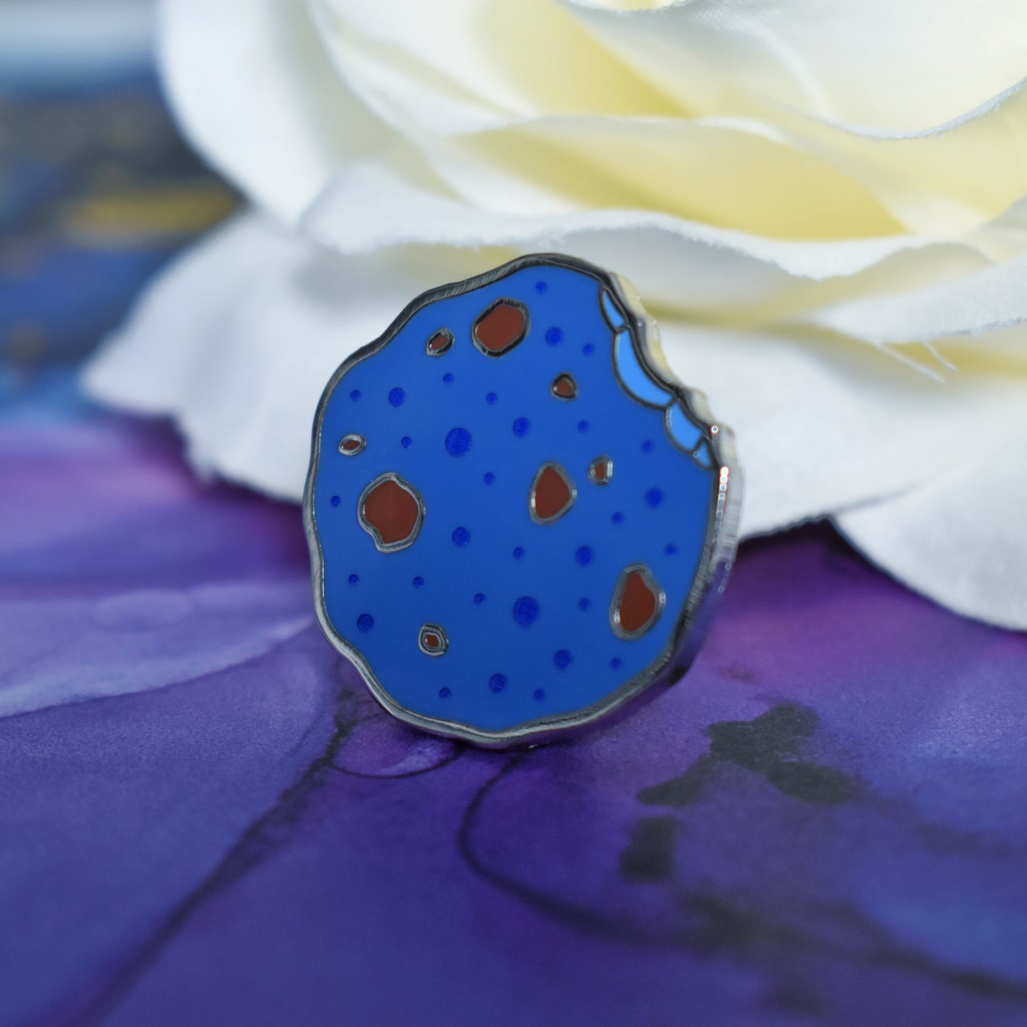 Silver metal enamel pin tilted to make the metal catch more light. Enamel pin is in a rough shape of a cookie but coloured blue, with darker blue spots and chunks of brown like it has chocolate in it. There is an edge that looks like a bite has been taken out of it. Background of photograph is blue/purple water inspired with a fake cream flower.