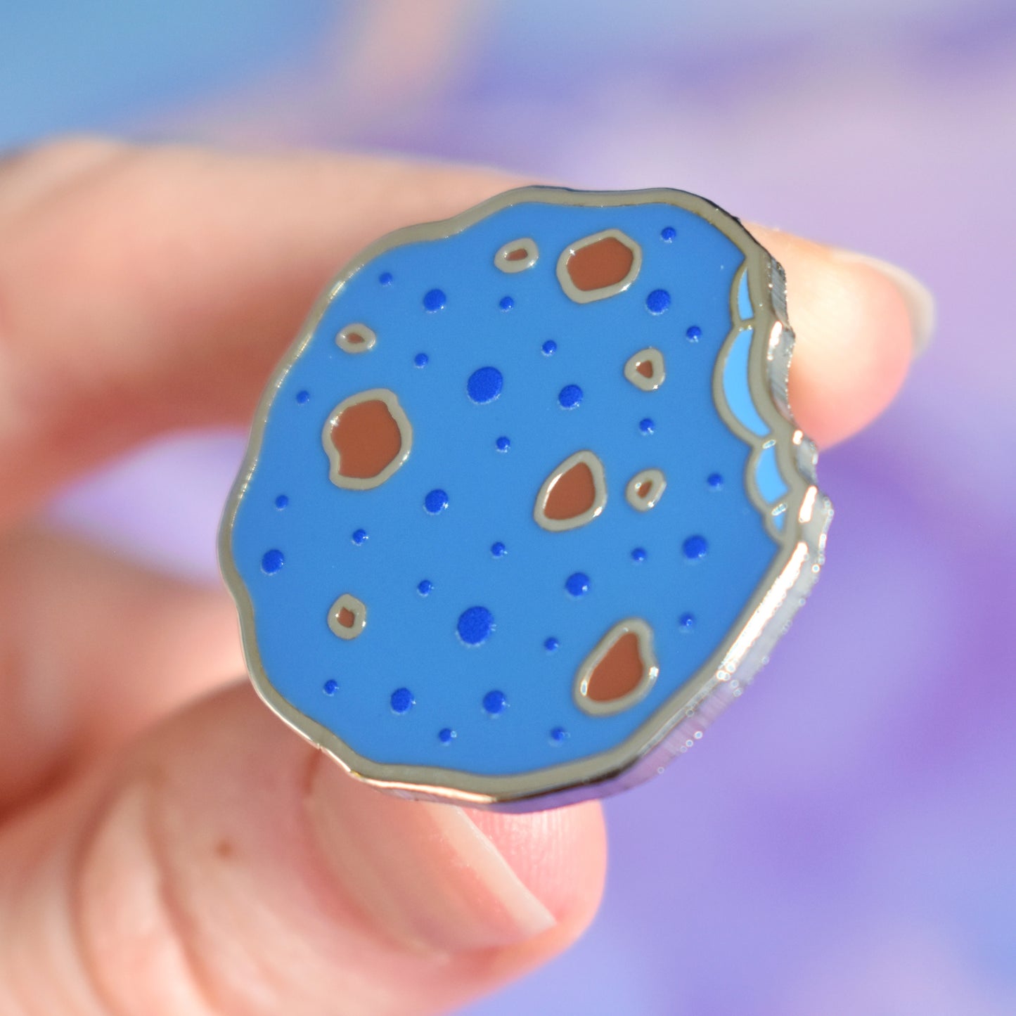 White hand holding silver metal enamel pin tilted to make the metal catch more light. Enamel pin is in a rough shape of a cookie but coloured blue, with darker blue spots and chunks of brown like it has chocolate in it. There is an edge that looks like a bite has been taken out of it. Background of photograph is blue/purple water inspired with a fake cream flower.