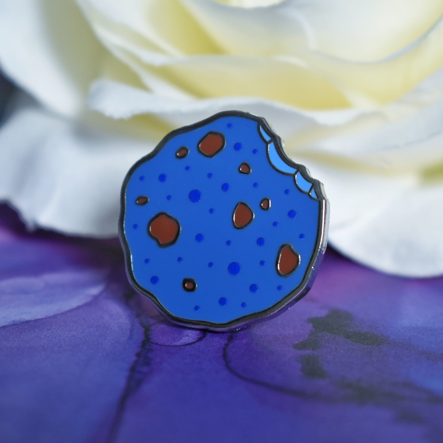 Silver metal enamel pin in centre of image. Enamel pin is in a rough shape of a cookie but coloured blue, with darker blue spots and chunks of brown like it has chocolate in it. There is an edge that looks like a bite has been taken out of it. Background of photograph is blue/purple water inspired with a fake cream flower.