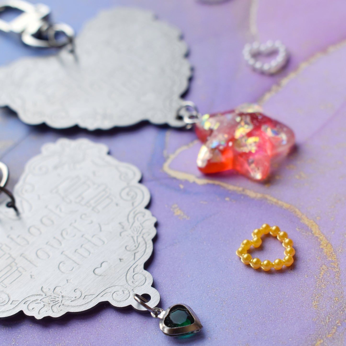 Close up of 2 keyrings with stainless steel engraved heart and book charm that reads book lovers club at diagonal in the centre of image. Left keyring has a small silver and gem dark green heart attached to charm. Right keyring has a pinky red glittery cabachon star attached to charm. On a pale blue/lilac watercolour background with different coloured heart beads scattered around.