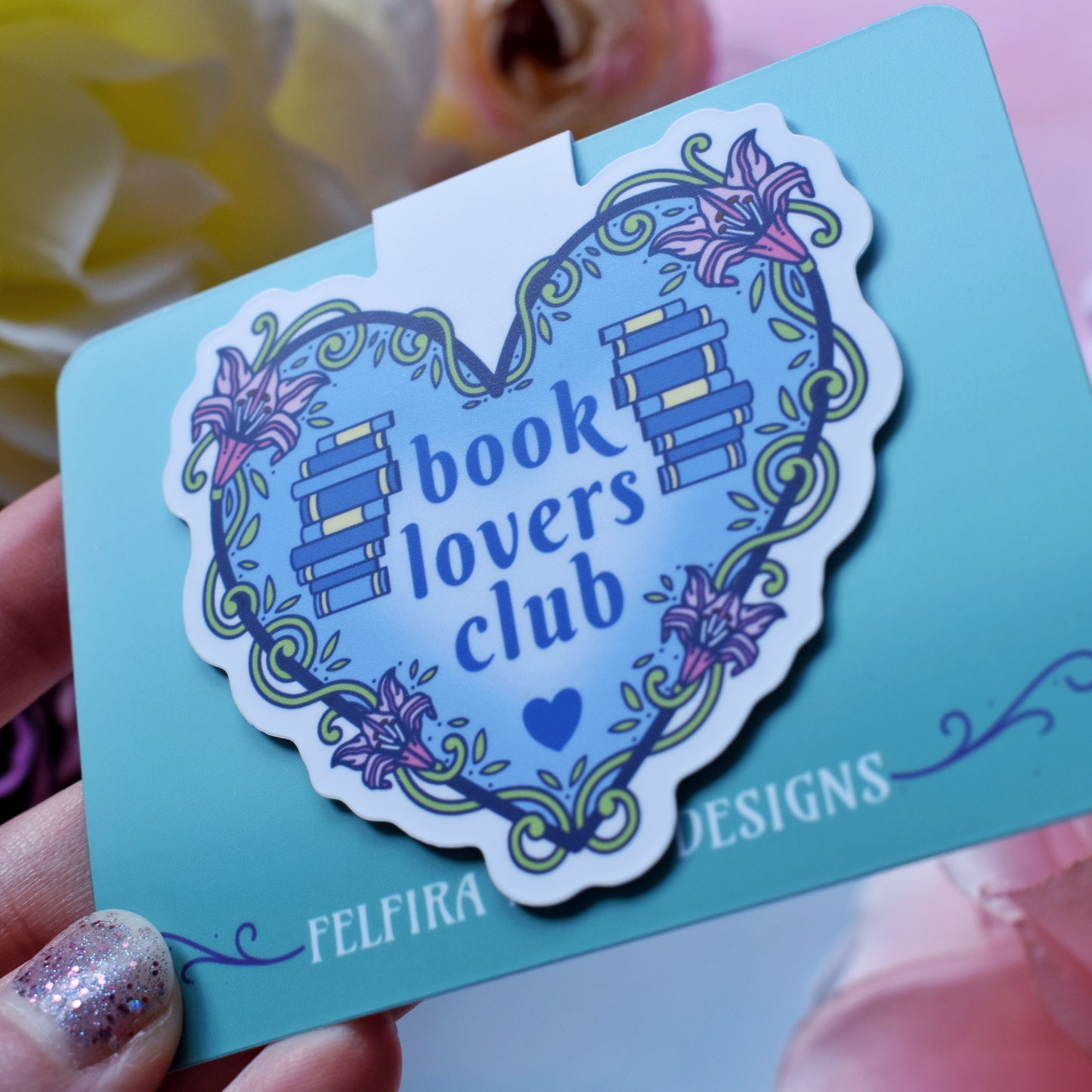 Blue heart-shaped design with a white border, featuring text 'book lovers club' with a floral and swirly outline with book spine stacks folded over a teal card. Card held by a white hand at an angle above a watercolour style background in blue, pink and purples with flowers around the edge of the photo.
