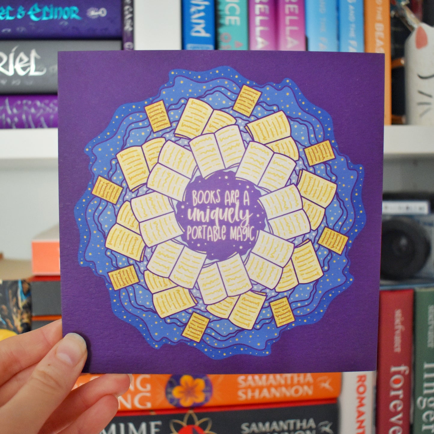 White hand holding print. Square print featuring a repeating pattern of yellow open books and blue swirls with purple around the edge. Design reads books are a uniquely portable magic in yellow at the centre of the design. Background of photograph has blurry bookshelf/books.
