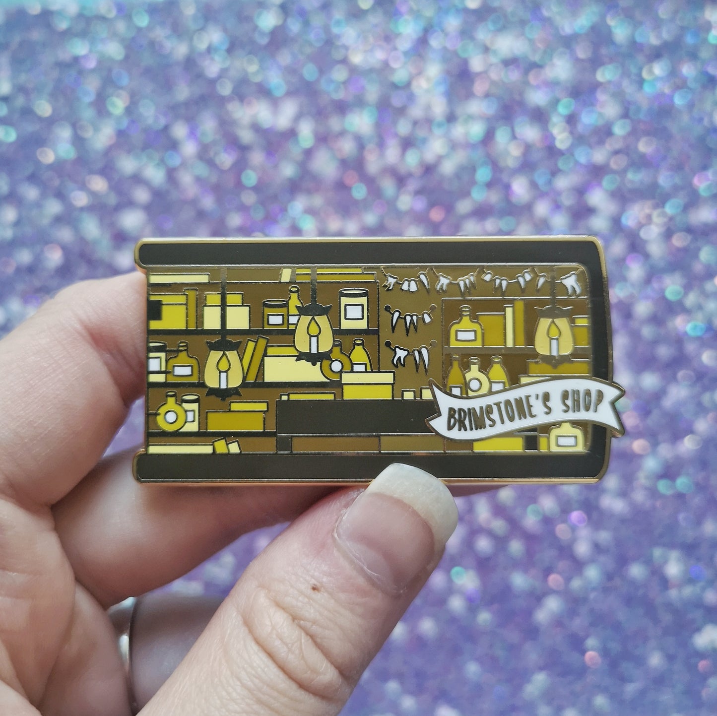 White hand holding gold metal enamel pin. Enamel pin is shaped like the end of a book in brown and features a scene in an old style shop/workshop. Lots of browns and yellows, candle lit lamps and teeth on the wall. A small banner reads brimstone's shop on the bottom right of the design. Background of photograph is sparkly blue/purple.
