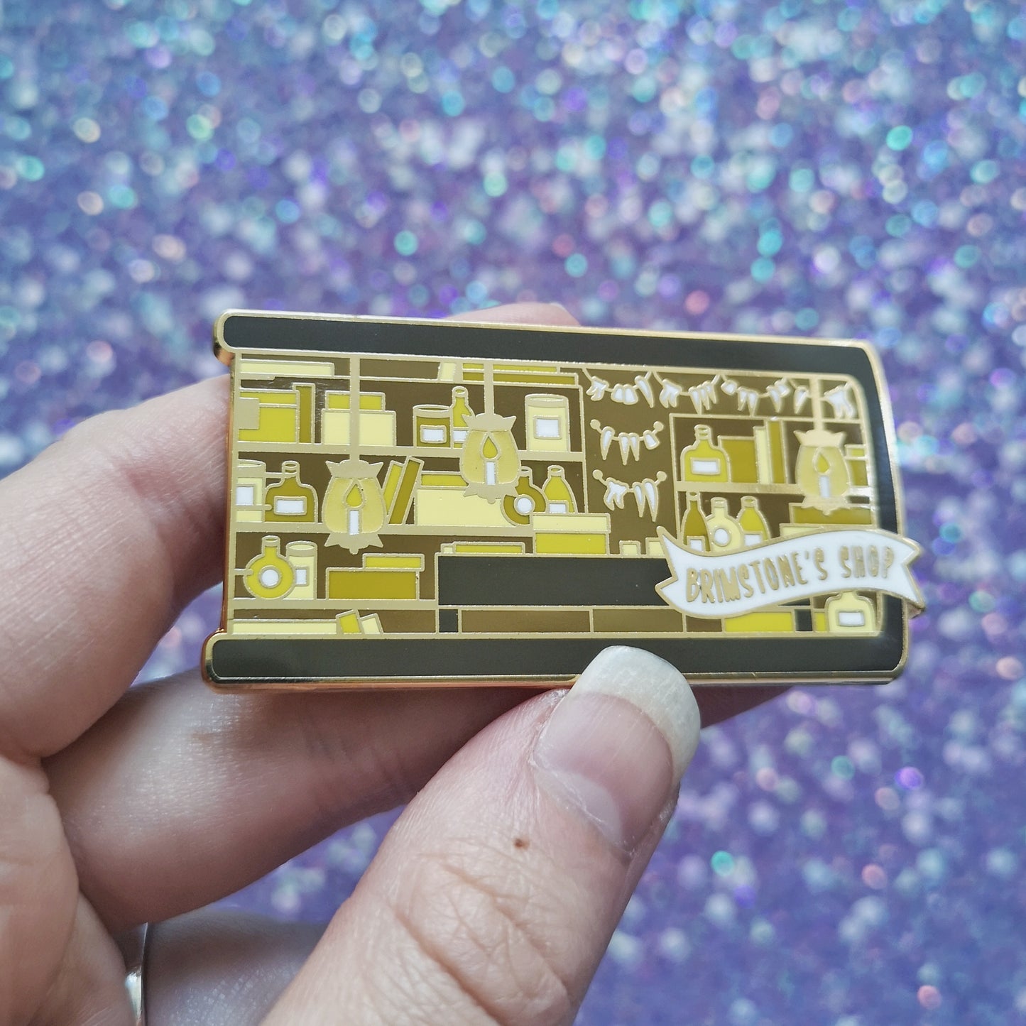 White hand holding gold metal enamel pin tilted to make the metal catch more light. Enamel pin is shaped like the end of a book in brown and features a scene in an old style shop/workshop. Lots of browns and yellows, candle lit lamps and teeth on the wall. A small banner reads brimstone's shop on the bottom right of the design. Background of photograph is sparkly blue/purple.