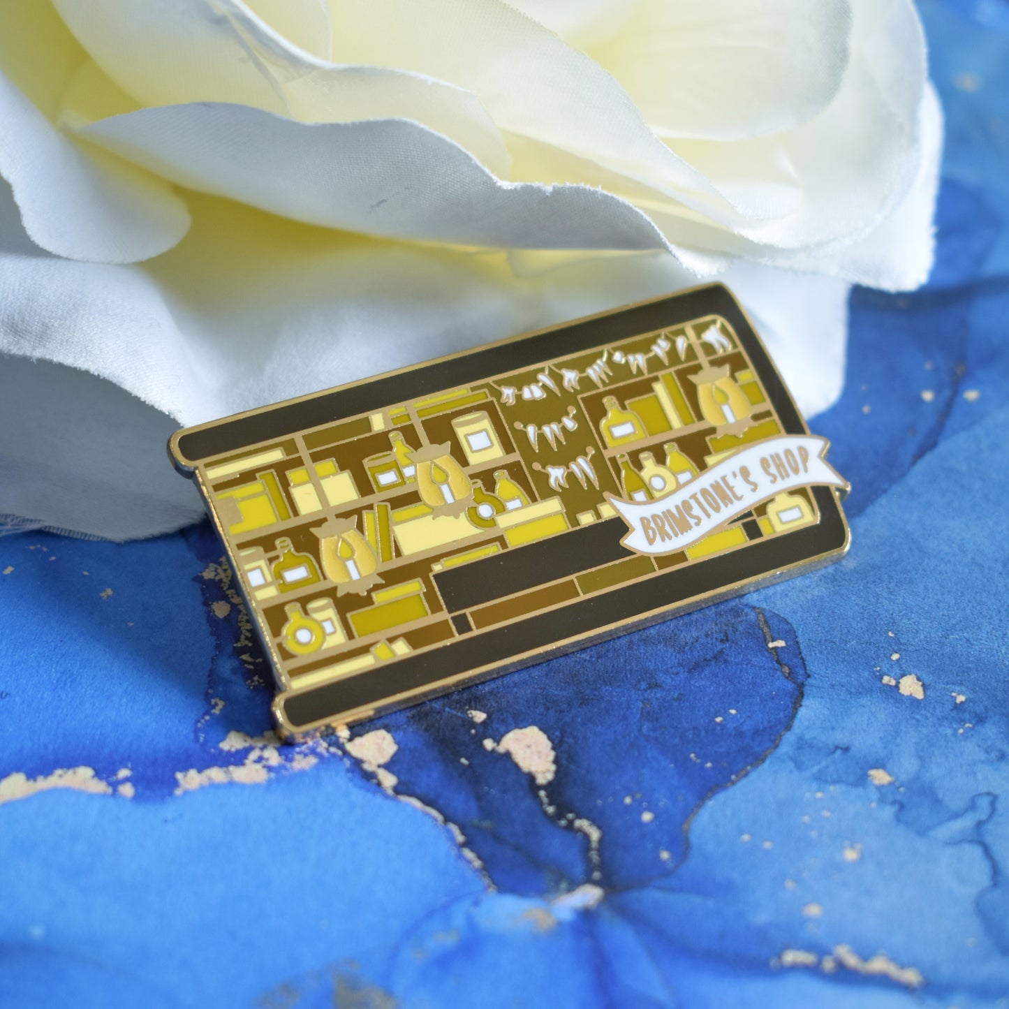 Gold metal enamel pin in centre of image. Enamel pin is shaped like the end of a book in brown and features a scene in an old style shop/workshop. Lots of browns and yellows, candle lit lamps and teeth on the wall. A small banner reads brimstone's shop on the bottom right of the design. Background of photograph is blue water inspired with a fake cream flower.
