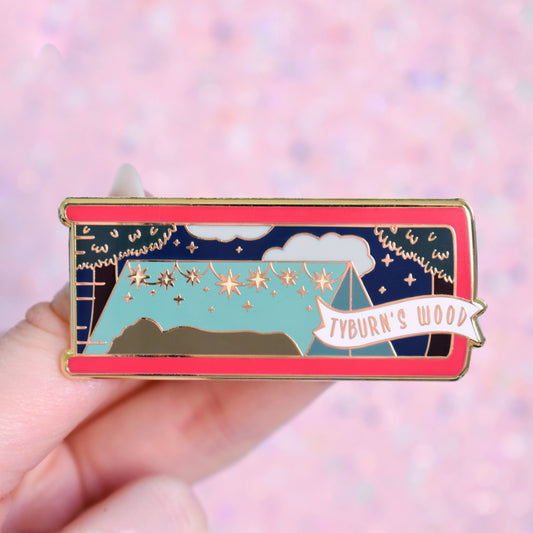 White hand holding gold metal enamel pin. Enamel pin is shaped like the end of a book in redish prink and features a scene with a text surrounded by trees and a night sky with clouds. The tent has the shape of people lay down and a string of fairy lights/sparkles. A small banner reads tyburn's wood on the bottom right of the design. Background of photograph is pink/peachy coloured.